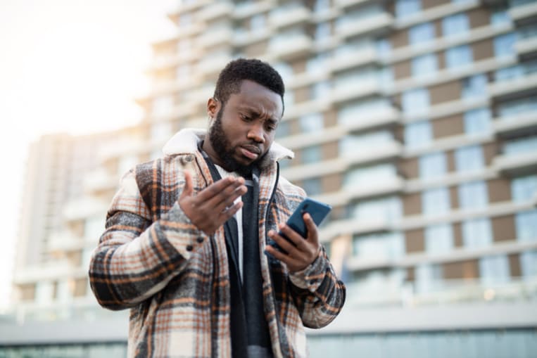 Young Black man stands outside and makes confused gesture while looking at his cell phone.
