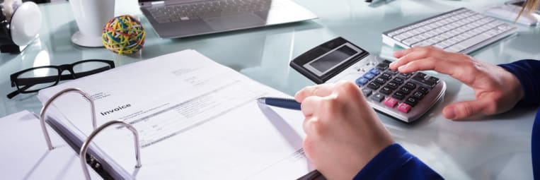 A person wearing a blue long-sleeve shirt is reviewing an invoice in a binder while using a calculator. Their right hand holds a pen, and their left hand presses buttons on the calculator. The workspace includes various items.